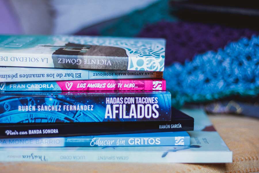 Torre de libros con los títulos a la vista en el lomo; de fondo, chal de ganchillo morado y turquesa