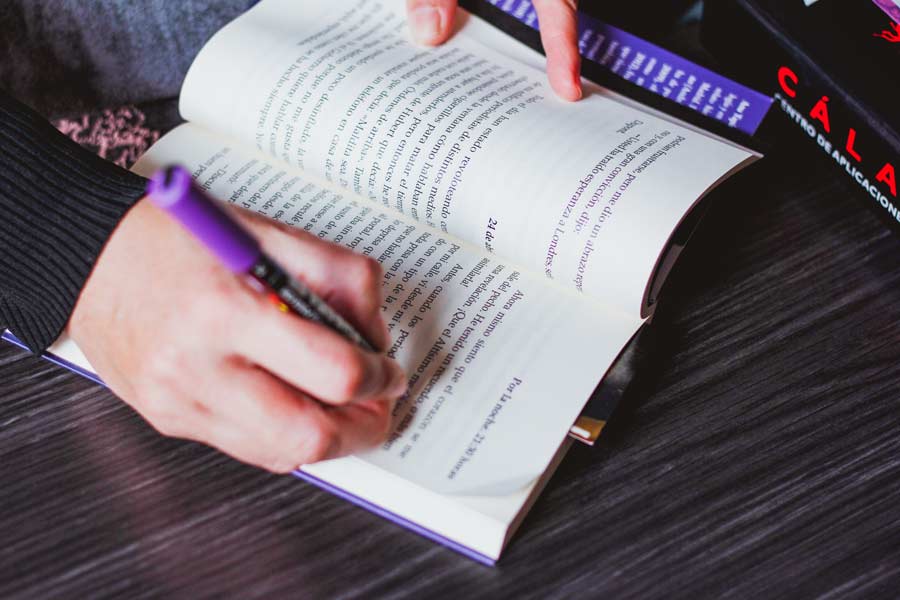 Libro abierto sobre una mesa; manos de M.ª Carmen sujetándolo y corrigiendo con un rotulador morado; fragmento de una carpeta de Cálamo & Cran.